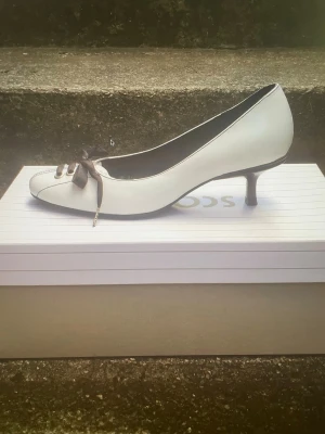 Vita pumps med svart rosett från Scorett - Säljer ett par vita pumps från Scorett med låg klack och rund tå. Skorna har svarta snören i rosett framtill och silverfärgade öljetter som ger en sportig touch. Tillverkade i ett slätt material med svarta detaljer längs kanten. Perfekta för dig som vill sticka ut!