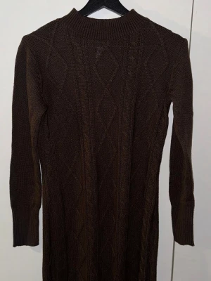 Stickad brun klänning från Rainbow - Mysig stickad klänning i mörkbrun färg från Rainbow. Klänningen har lång ärm, ribbad polokrage och ett snyggt kabelstickat mönster framtill. Perfekt för kyligare dagar och enkel att styla med boots eller sneakers.