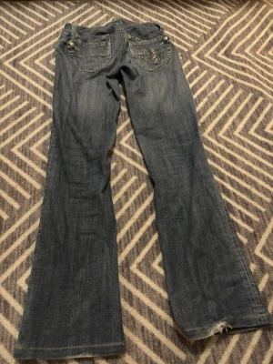 Blå bootcut jeans med nitar - Säljer ett par blå bootcut jeans med snygga nitar och mönster på bakfickan. Jeansen har klassisk femficksdesign, silverfärgade knappar och dragkedja. Materialet är jeans och passformen är loose med utsvängda ben. 