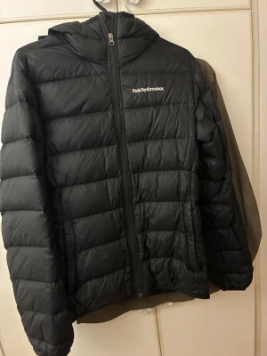Svart Peak Performance dunjacka S - Svart dunjacka från Peak Performance i storlek S. Jackan har huva, dragkedja framtill och ett quiltat mönster. Perfekt för vintern då den är varm och skön! Priset kan diskuteras!