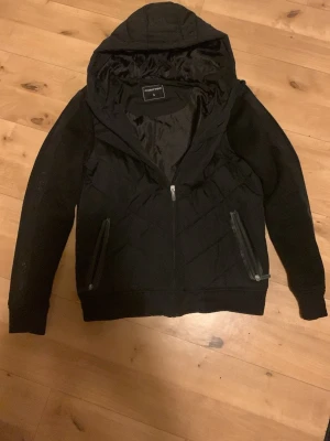 Svart pufferjacka från Chapter - Svart cardigan från Chapter, storlek S. Jackan har quiltad framsida och luva, dragkedja framtill och två fickor med dragkedja. Ärmarna är långärmade med en diskret textdetalj längs sidan. Perfekt för kyliga dagar.