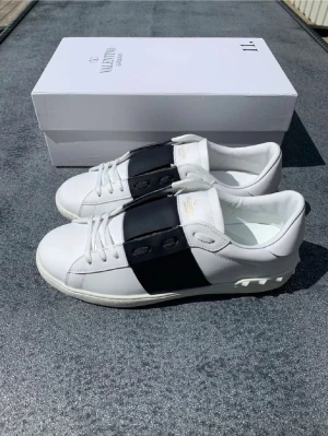 Valentino Garavani Open sneakers vit/svart - Snygga Valentino Garavani Open sneakers i vitt skinn med bred svart läderpanel över snörningen. Klassisk rund tå och platt sula med mönstrad undersida. Guldtext på plösen och vita skosnören. Kommer med originalkartong, dustbag och tags.