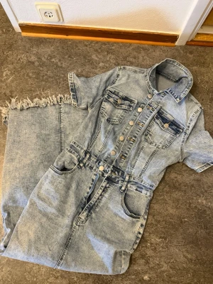 Ljusblå jeans jumpsuit med kort ärm - Säljer en ljusblå jeans jumpsuit med korta ärmar och fransad benslut. Den har flera fickor, tryckknappar framtill och klassisk krage. Materialet är jeans och den har en cool, avslappnad vibe. Perfekt för dig som gillar denim och vill sticka ut.
