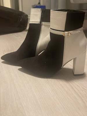 Svartvita stövletter med klack - Säljer ett par snygga stövletter i svart och vitt med spetsig tå och hög klack. Skorna har en cool textur på det vita partiet och ett dekorativt spänne med pärldetalj runt ankeln. Dragkedja på sidan för enkel på- och avtagning. Perfekt för dig som gillar statement boots. Kommer från SHEIN med original pris på 355 med tanke på att de är helt oanvända dock  finns små märken se bild 7 men dem har bara stått i ett skåp i något år då de inte passar mig leker syns inte då de är på sulan säljer för 340