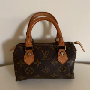 Louis Vuitton brun monogram handväska - Säljer en liten och klassisk handväska från Louis Vuitton med ikoniskt monogrammönster i brunt och beige. Väskan har två stadiga handtag i läder och en dragkedja upptill. Perfekt storlek för det viktigaste och en snygg detalj till din outfit.