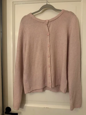 Ljusrosa stickad kofta med knappar - Mysig ljusrosa stickad kofta med rund hals och långa ärmar. Koftan har knappar framtill i matchande rosa färg och ribbade muddar vid ärmslut och nederkant. 