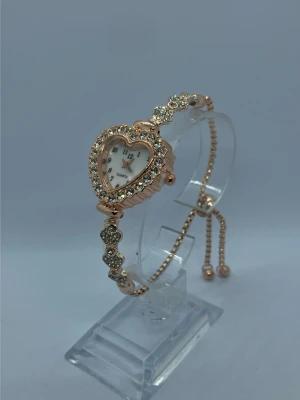Roséguld klocka med hjärtformad urtavla - Säljer en unik armbandsklocka i roséguld med hjärtformad urtavla. Klockan är dekorerad med glittrande stenar runt urtavlan och på armbandet. Justerbar kedja med små kulor i ändarna. Perfekt accessoar för dig som gillar detaljer och bling.rostfritt stål med ett nytt batteri 