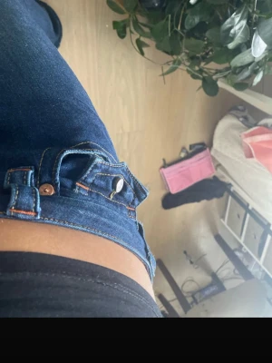 Blå bootcut jeans från True Religion - Säljer ett par mörkblå jeans från True Religion med klassisk bootcut och låg midja. Jeansen har tydliga orange sömmar, silverfärgade knappar och en ikonisk bakficka med logga. Materialet är jeans och passformen är loose. Perfekt för dig som gillar retrovibe och markerade detaljer. Finns synliga defekter som syns på bilderna.
