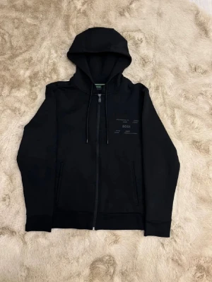  zip hoodie från BOSS - Svart hoodie från BOSS med stilren design, huva med snörning och hel dragkedja framtill. Tillverkad i mjuk bomull och har diskret BOSS-logga på bröstet. Två fickor fram och ribbade muddar vid ärmslut och nederkant. Perfekt för en chill och snygg look. Kom dm för eventuella frågor 🧐pris kan diskuteras💯. Har använt 1-5 gnr ny pris ligger på 2300.
