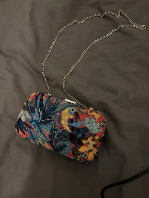 Färgglad kuvertväska med kedja - Säljer en unik kuvertväska med broderat blommönster i blått, rött, gult och svart. Väskan har en silverfärgad kedja och hårt skal, perfekt för att bära det viktigaste. Materialet är tyg med pärl- och paljettbroderier som ger en cool och färgstark look.