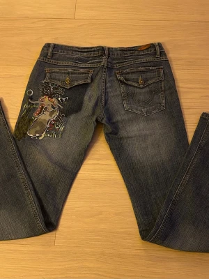 Blå jeans med broderad konst på bakfickan - Unika blå jeans med snygg broderad konst på ena bakfickan. Modellen har klassisk femficksdesign, raka ben och knäppning med knapp och dragkedja. Materialet är slitstarkt jeans och färgen är mörkblå med lätt tvättad look. Lite slitna nedtill som bara ser coolt ut. Storlek W 29.