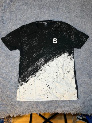 Svartvit t-shirt Black Squad XL - Säljer en t-shirt från Black Squad i storlek XL. T-shirten är svart och vit med coolt splattermönster och en stor B-logga på bröstet. Materialet är 100% bomull och modellen har korta ärmar. Perfekt för dig som gillar streetwear och vill sticka ut.