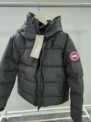 Svart dunjacka från Canada Goose M - Svart dunjacka från Canada Goose med huva och ikonisk röd-vit-blå patch på ärmen. Jackan har dragkedja, knappar och snörning vid huvan. Insidan har en praktisk ficka med dragkedja. Perfekt för kalla dagar och riktigt snygg streetstil.