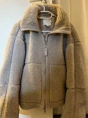 Beige teddyjacka med dragkedja - Mysig beige teddyjacka med hög krage och hel dragkedja framtill. Jackan har två stora fickor och är tillverkad i fluffigt fleece-material som ger en skön och varm känsla. Perfekt för kyliga dagar och har en loose passform för extra komfort.