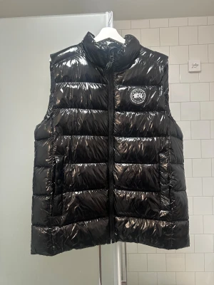 Canada Goose väst  - Säljer en svart, glansig dunväst från Canada Goose med hög krage och broderad logotyp på bröstet. Västen har quiltad design, dragkedja framtill och praktiska fickor med knappstängning. Perfekt för kyliga dagar och snygg att styla över hoodie eller tröja. Storlek: L