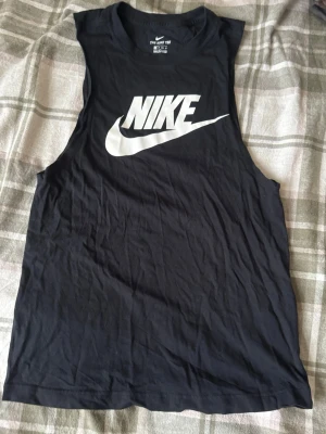 Svart linnetopp från Nike XS - Svart linnetopp från Nike i storlek XS med stort vitt Nike-logotryck på bröstet. Modellen är ärmlös med rund halsringning och tillverkad i mjuk bomull, perfekt för träning eller chill. Snygg och sportig design som är enkel att matcha.