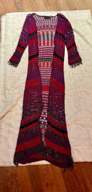 Lång kofta från Karen Millen i lila och rött - Säljer en festlig , glittrig , lång, öppen kofta från Karen Millen med färgglada ränder i lila, rött, svart och vitt. Koftan är stickad med glittriga detaljer, har fransar vid ärmslut och nederkant samt knappar framtill. Perfekt för dig som vill sticka ut med en unik och bohemisk look. Karen Millen storlek 2 är small tror jag men passar M också