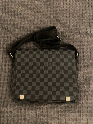 rutig axelväska i svart - Säljer en snygg axelväska med klassiskt rutigt mönster i svart och grått. Väskan har justerbar axelrem och silverfärgade detaljer. Perfekt storlek för att få plats med allt viktigt och har en stilren design som sticker ut.