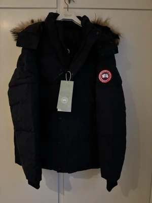 Mörkblå dunjacka från Canada Goose - Säljer en mörblå dunjacka från Canada Goose med ikonisk röd patch på ärmen och  huva med snygg pälsdetalj som går att ta av. Jackan har dragkedja och knappar framtill, quiltad design och långärmade ärmar. Perfekt för kalla vinterdagar och riktigt stilren look.