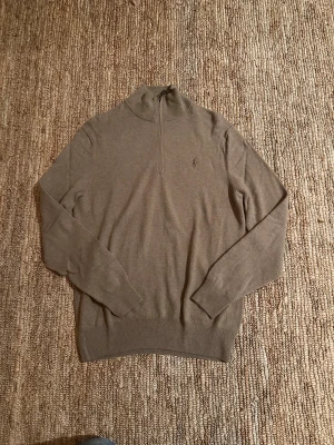 Beige quarterzip från Polo Ralph Lauren - Snygg beige quarterzip från Polo Ralph Lauren med diskret broderad logga på bröstet. Tröjan har half zip i halsen, ribbade muddar och är långärmad. Perfekt för dig som gillar klassisk stil med en modern touch.