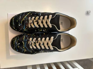 Maison Margiela svart sneakers med färgstänk - Maison Margiela sneakers i svart skinn med unikt färgstänksmönster. Beige snörning och bruna platta gummisulor. Rund tå och låg profil. Perfekt för dig som gillar statement och konstnärlig vibe. Prisförslag och frågor är det bara att skicka. Även byte mot andra skor är intressant 🧐🤯👑