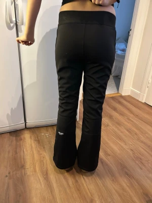 Svarta skidbyxor från 8848 Altitude - Svarta skidbyxor från 8848 Altitude med bootcut-snitt och ribbade detaljer vid knäna. Byxorna har dragkedja och dubbla knappar i midjan, samt elastisk midja för extra komfort. Perfekta för vintern och gjorda i slitstarkt syntetmaterial.