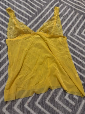 Gul spetsdetaljerad linnetopp - Säljer en tunn och transparent gul linnetopp med spetsdetaljer och blommönster vid axelbanden. Toppen har v-ringad hals och ärmlös design med smala axelband. Märket och storleken står inte men skulle säga att det är en xs/s