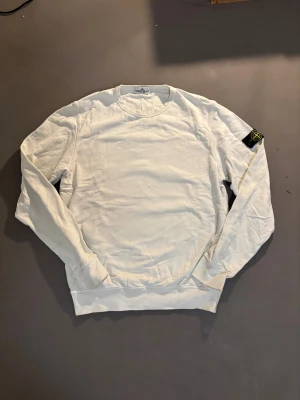 Vit sweatshirt från Stone Island Junior - Vit sweatshirt från Stone Island Junior med klassisk rund hals och långärmad passform. Tröjan har den ikoniska Stone Island-patchen på vänster ärm och är tillverkad i mjuk bomull. Perfekt för dig som gillar clean och stilren streetwear.