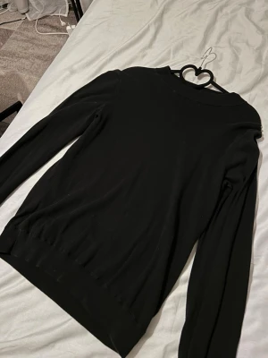 Svart långärmad tröja från Zara och Riley - En stilren svart långärmad tröja från Zara och Riley med rund hals och ribbade muddar vid ärmslut och nederkant. Tröjan har en klassisk passform och är tillverkad i mjukt bomullsmaterial som känns skönt mot huden. Perfekt för lager-på-lager under hösten och vintern.