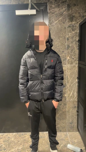 Svart pufferjacka från Ralph Lauren - Säljer en svart pufferjacka från Ralph Lauren med huva och dragkedja framtill. Jackan har en röd broderad logga på bröstet och är vadderad för extra värme. Perfekt för kalla dagar och har en clean, sportig look.