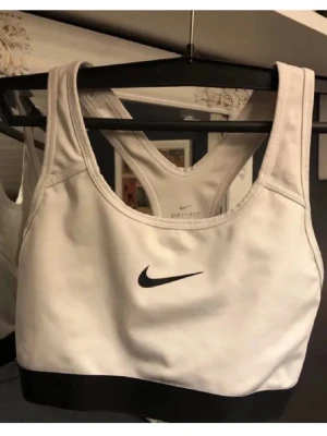 Vit sport-bh från Nike Dri-Fit - Säljer en vit sport-bh från Nike med svart logga framtill och svart kant nedtill. Modellen är Dri-Fit och har brottarrygg för extra stöd vid träning. Materialet är syntet och designen är stilren och sportig.