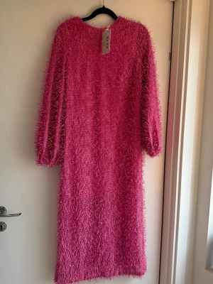 Rosa fransig långklänning från NA-KD - Säljer en maxiklänning från NA-KD i stark rosa med fransig textur och lång ärm. Klänningen har en djup öppen rygg och är tillverkad i syntetmaterial. Den har en cool, unik look som verkligen sticker ut.