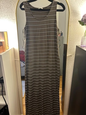 Randig långklänning i bomull - Säljer en snygg långklänning med bruna och vita horisontella ränder. Klänningen är ärmlös och har en rund halsringning. Materialet är mjuk bomull, perfekt för varma dagar. Enkel och stilren design som är lätt att matcha med accessoarer.