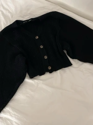 Svart stickad kofta med knappar - Säljer en svart stickad kofta med v-ringad hals och fyra snygga knappar framtill. Koftan är croppad och har långa, lite bredare ärmar med ribbade muddar. Perfekt att slänga över en t-shirt eller linne för en chill look.