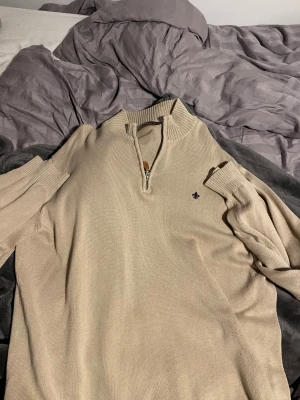 Beige stickad half zip tröja Morris - Snygg beige stickad tröja från Morris i storlek XXL. Tröjan har en ribbad krage och half zip-dragkedja framtill. Liten broderad logga på bröstet och långa ärmar med ribbade muddar. Perfekt för dig som gillar klassisk och stilren design.