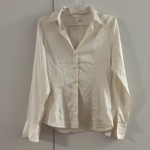 Krämvit figurnära skjorta från H&M - Säljer en stilren krämvit silkes skjorta från H&M i storlek S. Skjortan har klassisk krage, lång ärm med bred manschett och knappar framtill. Materialet är mjukt och har en lätt glansig finish som ger en lyxig känsla. Perfekt för dig som gillar enkel och clean stil.