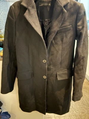 Svart kavaj från Zara Woman - Stilren svart kavaj från Zara Woman med klassisk krage, två knappar framtill och fickor med lock. Lång modell som ger en snygg siluett och funkar perfekt till många olika looks. Fodrad insida för extra komfort.