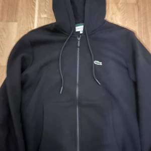 Äkta Lacoste kofta med kvitto - Svart hoodie från Lacoste i klassisk passform, med huva och snörning. Tröjan har dragkedja framtill och den ikoniska gröna krokodilloggan broderad på bröstet. Mjuk bomullskänsla och praktisk ficka framtill. Perfekt för chill dagar.
