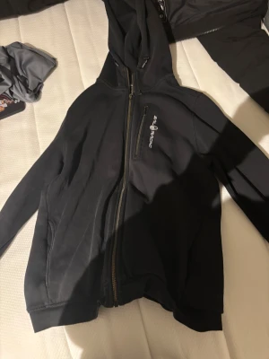 Svart hoodiejacka från Sail Racing - Svart hoodiejacka från Sail Racing med dragkedja hela vägen och logga på bröstet. Jackan har en praktisk bröstficka med dragkedja och huva med justerbara snören. Perfekt för chill dagar och streetwear-stil.