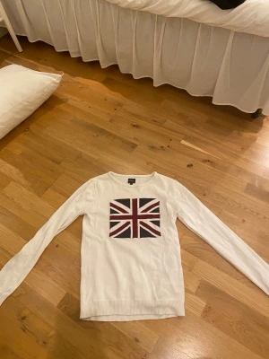 Vit stickad tröja med Union Jack från Solid - Vit stickad tröja från Solid i storlek M med långärmad passform och ribbade muddar. På bröstet finns ett stort Union Jack-tryck i marinblått och vinrött. Rund halsringning och mjukt material.