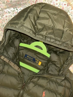 Olivgrön pufferjacka från Polo Ralph Lauren - PRISET GÅR ATT DISKUTERA Säljer en olivgrön pufferjacka från Polo Ralph Lauren med huva och dragkedja framtill. Jackan har två sidofickor med dragkedja och den klassiska Polo-loggan broderad på bröstet. Materialet är syntet och jackan har en quiltad design som ger en schysst look.