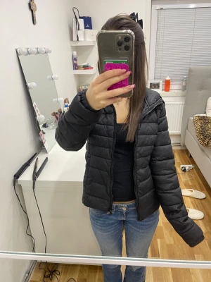 Svart  tunn pufferjacka med huva - Säljer en svart pufferjacka med huva och dragkedja framtill. Jackan har ett quiltat mönster och är i syntetmaterial, perfekt för mindre kyliga dagar. Passformen är normal och jackan har långa ärmar. Enkel och stilren design som funkar till jeans eller andra outfits.