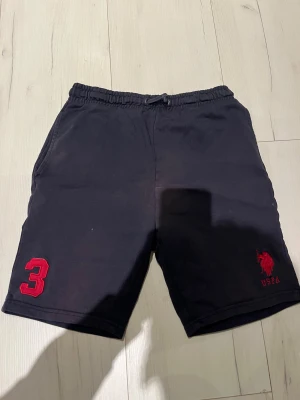 Svarta shorts från US Polo Assn - Svarta shorts från US Polo Assn med röd broderad logga och siffra 3 på ena benet. Mjuk bomullskänsla, elastisk midja med snörning och fickor på sidorna samt en bakficka. Perfekta för chill dagar eller sport.