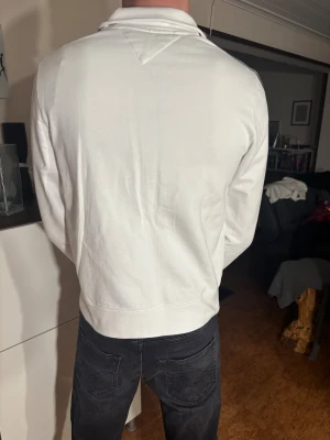 Tommy Hilfiger helzip tröja - Snygg vit tröja från Tommy Hilfiger med dragkedja och två fickor fram. Längs ärmarna går stilrena ränder i marinblått och vinrött. Tröjan har hög krage och logga på bröstet. Perfekt för dig som gillar sportig och clean stil. Modellen är 190 cm lång och väger 70kg