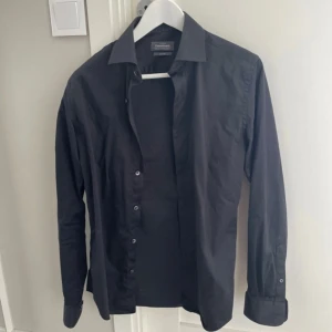 Svart skjorta från Dressmann - Stilren svart skjorta från Dressmann med klassisk krage och knappar framtill. Skjortan har lång ärm och är tillverkad i mjuk bomull som känns skön mot huden. Perfekt för dig som gillar enkel och snygg stil.