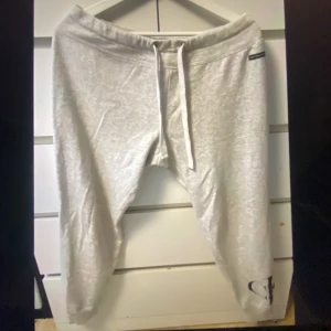 Ljusgrå mjukisbyxor med snörning från Calvin Klein  - Säljer ett par ljusgrå mjukisbyxor från Calvin Klein i storlek S med snörning i midjan och en liten CK logga vid benet. Byxorna är tillverkade av 60% bomull och 40% återvunnen polyester, vilket gör dem både mjuka och hållbara. Perfekta för vilo dagar hemma.