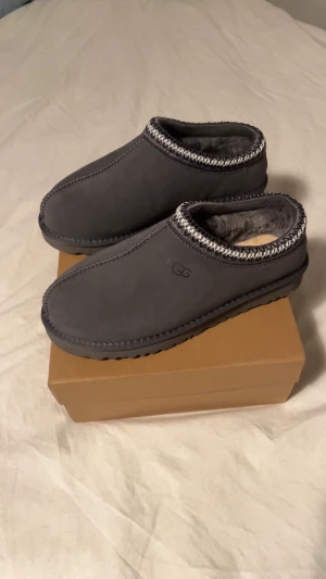 Grå UGG slip-in mockatofflor - Säljer ett par grå slip-in tofflor från UGG med mjuk päls på insidan och snyggt mönstrad kant. Yttermaterialet är mocka och sulan är robust med UGG-logga undertill. Perfekt för att hålla fötterna varma och sköna under kyliga dagar.