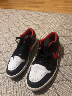 Nike Air Jordan 1 Low svart/röd - Nike Air Jordan 1 Low sneakers med svart och vitt skinn, röda detaljer och röd swoosh. Låg modell med perforerad vit tå, svart snörning och röd insida. Ikonisk vinge-logga på hälen och klassisk platt sula. Perfekt för dig som gillar streetstyle och sneakers med attityd.
