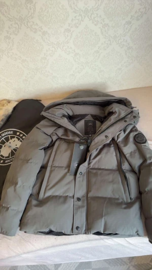 Grå wyndham dunjacka från Canada Goose - Snygg grå dunjacka från Canada Goose med svart patch på ärmen och avtagbar huva med fuskpäls. Jackan har dragkedja, knappar och två stora fickor framtill. Perfekt för kalla vinterdagar och riktigt stilren look.