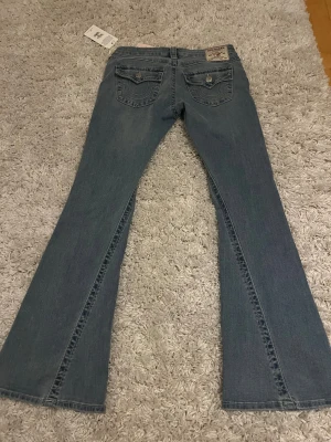 True Religion Joey Low Rise Flare Jeans - Snygga blå jeans från True Religion, modell Joey Low Rise Flare. De har låg midja, utsvängda ben och klassiska detaljer som markerade sömmar och fickor med lock och knappar bak. Perfekta för dig som gillar en retroinspirerad look.